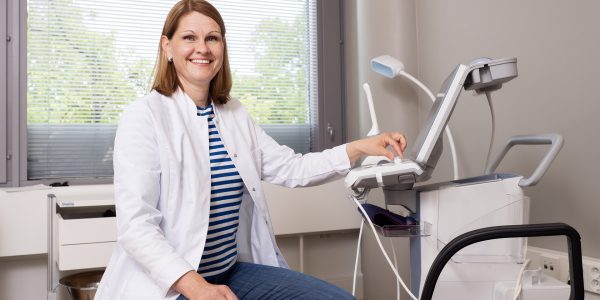 Naistentautien ja synnytyksen erikoislääkäri istumassa ja hymyilemässä tuolilla vastaanottohuoneessaan käsi ultraäänilaitteen säätimellä.