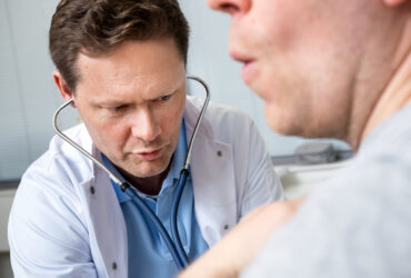 Työterveyslääkäri kumartuu potilaan puoleen ja tutkii stetoskooppi korvillaan kuvassa etualalla osittain näkyvää asiakasta.